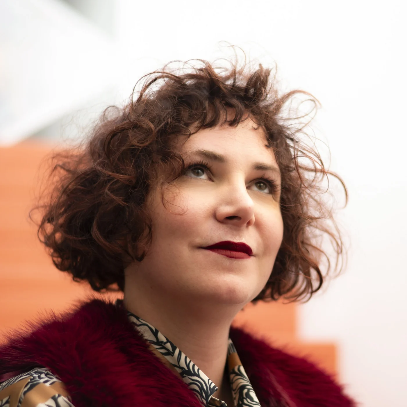 Alice Kielo, portrait: curly hair, eyes raised, burgundy fur jacket